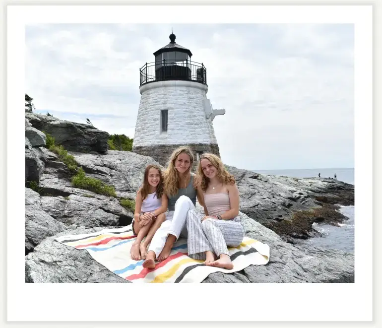 Three women sitting on a blanket near a lighthouse on rocky terrain.