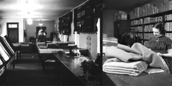 Black and white photo of a person in a room with stacked towels and a cluttered table.