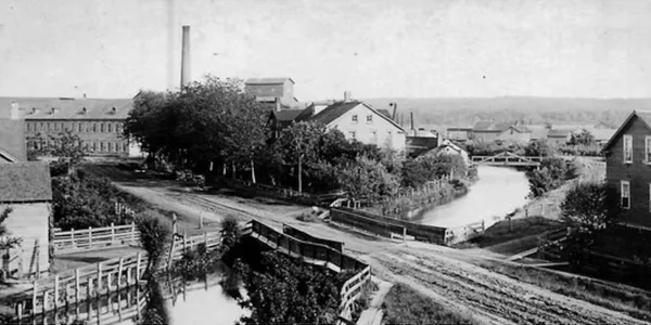 Historic black and white photo of a riverside village with a bridge and railway tracks.