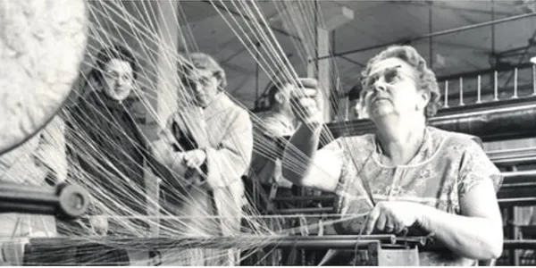 Three elderly women weaving threads on a loom in a workshop.