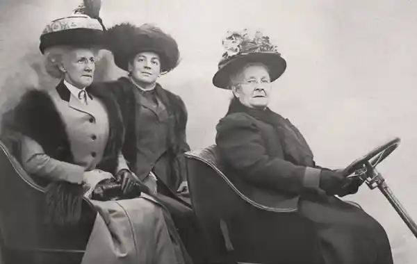 Three women from the early 1900s wearing large hats, sitting in a carriage.