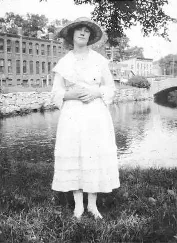 Young woman in early 20th-century dress standing by a river.