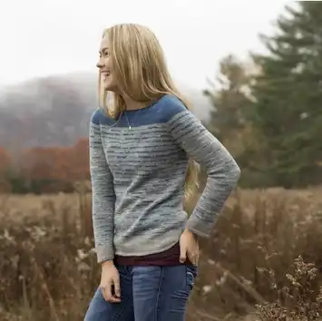 A woman stands outdoors in a gray and blue sweater, looking to the side.
