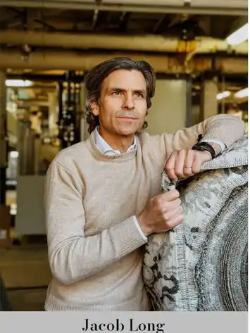 Man in a beige sweater standing beside a stone sculpture indoors.