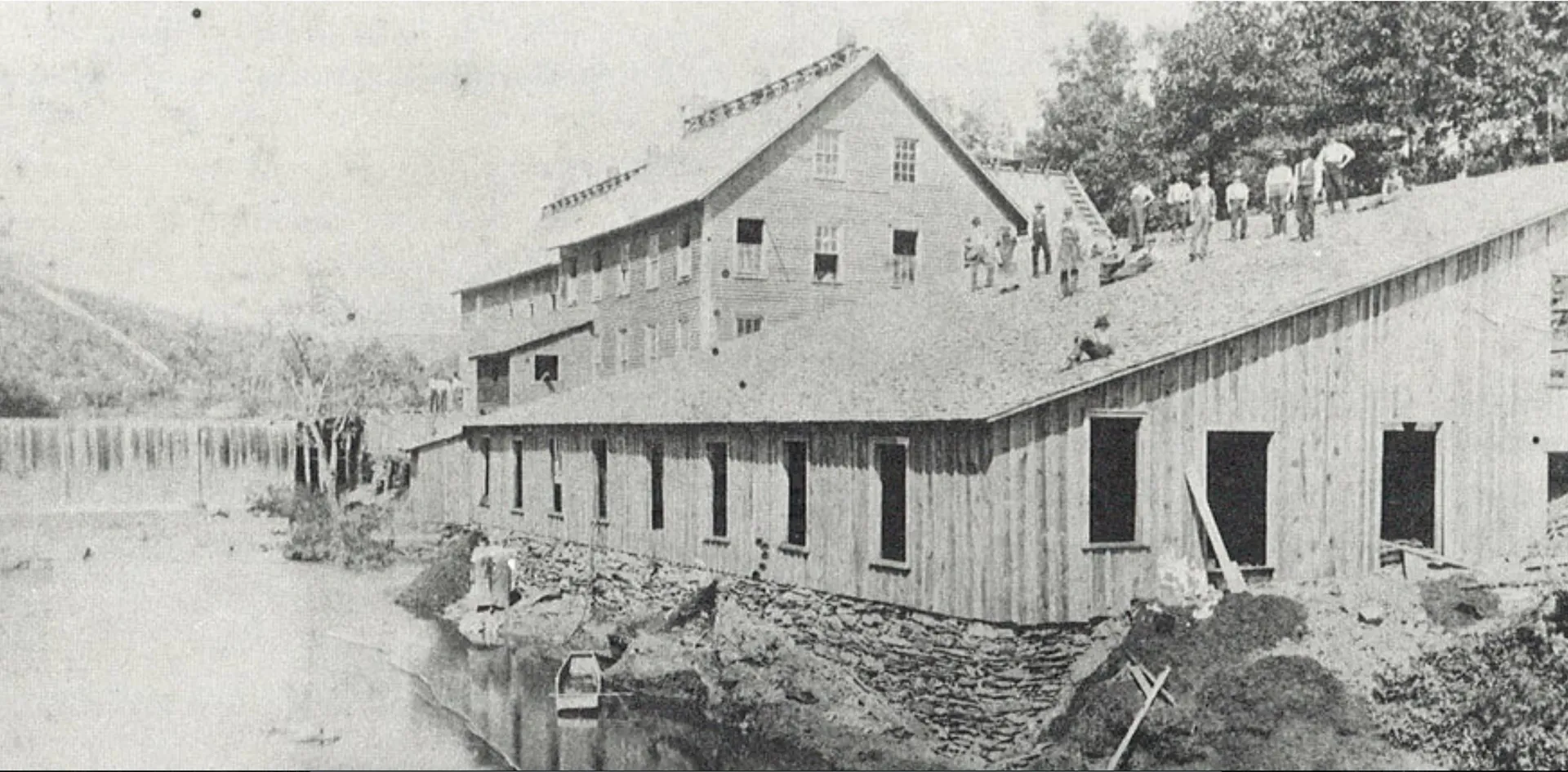Black-and-white image of an old wooden industrial building with a creek in front.