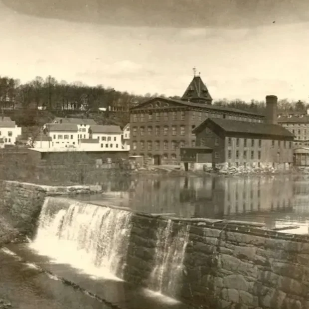 Old sepia photo of a waterfall with buildings and a church steeple in the background.